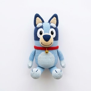 May include: A handmade crochet dog toy in shades of blue, with a red collar and a small gold bell. The dog has a light tan muzzle and a friendly expression. The toy is sitting upright, showcasing its detailed craftsmanship.