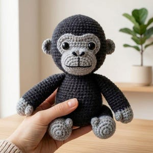 May include: A handmade crochet gorilla doll in shades of gray and black. The plush toy features a detailed face with large eyes, a smiling mouth, and small ears. The gorilla is being held by a person.