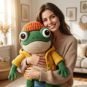 May include: A large, handmade crochet frog doll with a green body, orange beanie, and yellow jacket. The frog has large, expressive eyes and is being held by a person. The doll is a whimsical and charming handmade item.