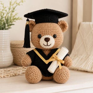 May include: A crocheted teddy bear wearing a black graduation cap with a gold tassel and a black and gold graduation gown. The bear holds a rolled-up diploma tied with a gold ribbon. The bear is light brown with black eyes and a white muzzle.