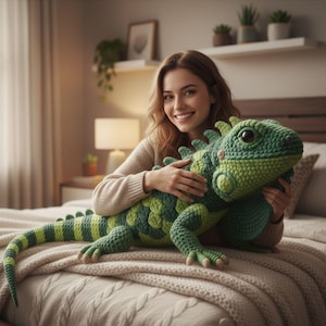 Op de afbeelding: Een grote, handgemaakte gehaakte leguaan in groen- en geeltinten. Het pluche reptiel heeft gedetailleerde schubben, klauwen en een lange staart. De leguaan ligt op een bed met een beige deken.