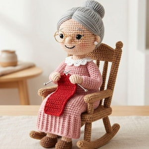 May include: A crocheted doll of a grandmother figure, seated in a wooden rocking chair. The doll wears a pink dress, glasses, and a bun hairstyle. She is holding knitting needles and a red scarf. The doll has a smiling expression.
