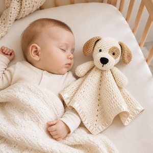 May include: A cream-coloured crochet security blanket featuring a stuffed dog head with brown ears and a brown eye patch. The blanket has a textured square body with a smooth satin border trim, designed as a soft comfort object for infants.