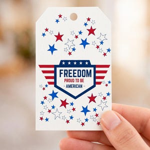 May include: A white rectangular gift tag with a hole at the top, featuring a central blue shield emblem with red horizontal stripes and the text FREEDOM PROUD TO BE - AMERICAN. The tag is decorated with scattered red, blue, and outlined stars.