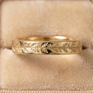 May include: A gold-colored ring with a detailed leaf pattern. The ring is set against a beige velvet background inside a ring box. The band features an engraved design of leaves and vines, creating a nature-inspired aesthetic.