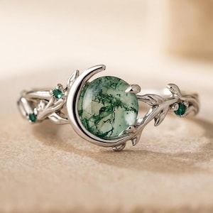 May include: A silver-toned ring featuring a round, translucent moss agate stone with green inclusions. The band is designed with a crescent moon shape partially framing the stone, accented by small green gemstones and a textured, branch-like motif.