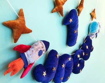 May include: A felt banner with a space theme, featuring a rocket, stars, and the name "JAKE" in blue with gold stars. A large blue number "1" is also included, along with a planet. The banner is strung with white string and has a light blue background.