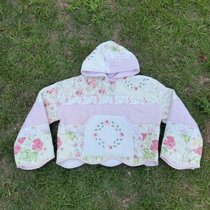 Puede incluir: Un suéter acolchado con capucha confeccionado en tela de patchwork que presenta una mezcla de patrones florales en rosa y blanco, cuadros vichy y paneles en color crema. La prenda incluye un bolsillo tipo canguro con un bordado de corona floral.