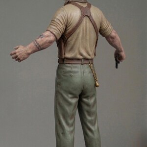 May include: A miniature figurine of a person with short hair, wearing a tan short-sleeved shirt, brown leather braces, and olive green trousers. The figure has a tattoo on the right arm, a gold chain hanging from the belt, and brown boots.