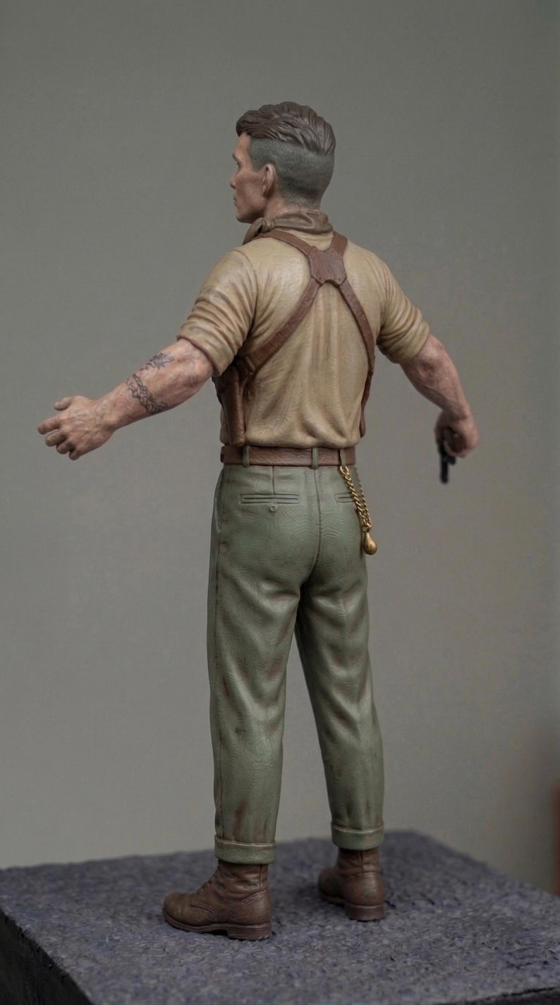 May include: A miniature figurine of a person with short hair, wearing a tan short-sleeved shirt, brown leather braces, and olive green trousers. The figure has a tattoo on the right arm, a gold chain hanging from the belt, and brown boots.
