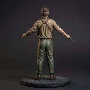May include: A painted figurine of a person viewed from behind, wearing a tan short-sleeved shirt, olive green trousers, and a brown leather shoulder holster. The figure has short brown hair and holds a small metallic handgun in the left hand.