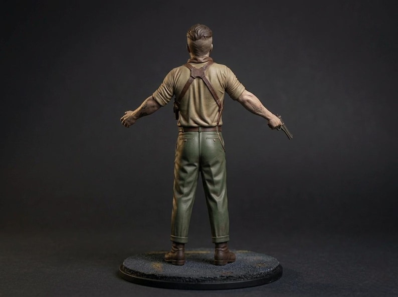 May include: A painted figurine of a person viewed from behind, wearing a tan short-sleeved shirt, olive green trousers, and a brown leather shoulder holster. The figure has short brown hair and holds a small metallic handgun in the left hand.
