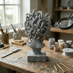 Puede incluir: Una escultura gris de una cabeza con numerosas serpientes formando el cabello, sobre una base cuadrada con un peque&ntilde;o s&iacute;mbolo grabado en el frente. El busto presenta escamas detalladas en la zona del cuello y se sit&uacute;a sobre un banco de trabajo.