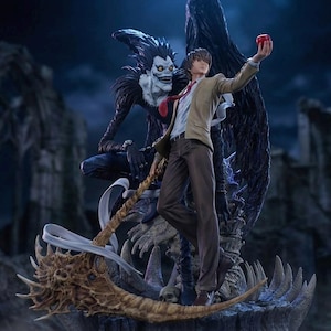 Può includere: Statua dettagliata con due personaggi dell'anime Death Note. Una figura tiene in mano una mela rossa, mentre l'altra, uno Shinigami, ha grandi ali nere. Le figure sono ambientate su uno sfondo scuro e gotico.