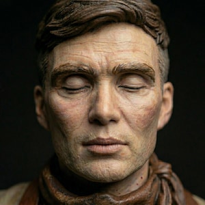 May include: A highly detailed, realistic sculpture of a person's head and shoulders. The figure has short, dark brown hair, closed eyes, and a neutral expression. A textured, dark brown scarf is wrapped around the neck against a black background.