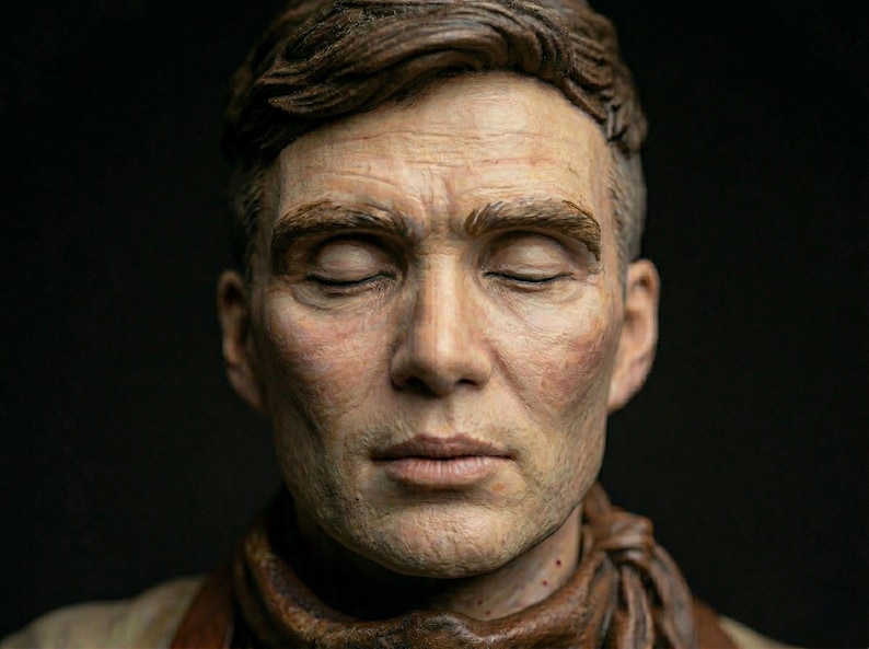 May include: A highly detailed, realistic sculpture of a person's head and shoulders. The figure has short, dark brown hair, closed eyes, and a neutral expression. A textured, dark brown scarf is wrapped around the neck against a black background.