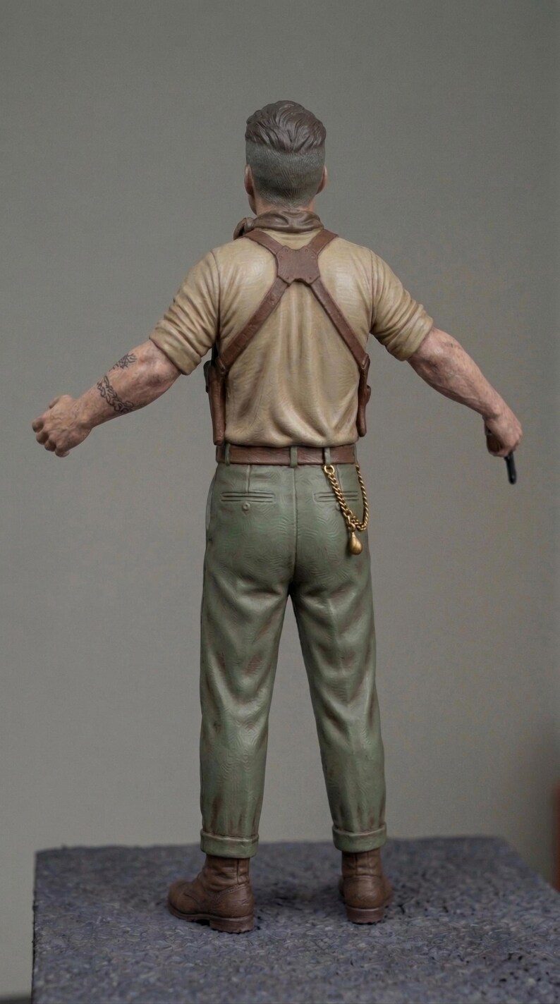 May include: A miniature figurine of a person seen from the back, wearing a tan short-sleeved shirt, olive green trousers, and brown boots. The figure features a brown leather shoulder holster, a gold-coloured pocket watch chain, and a tattoo on the right arm.