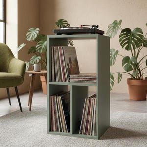 Op de afbeelding: Een saliegroene houten opbergkast met drie open vakken voor vinylplaten. Op het bovenoppervlak staat een zwarte platenspeler. De kast heeft één grote bovenplank en twee kleinere planken naast elkaar onderin, allemaal gevuld met vinylplaten.