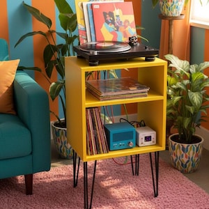 Puede incluir: Un soporte amarillo para tocadiscos con patas de horquilla negras. Un tocadiscos negro se encuentra en la parte superior, con discos de vinilo y un componente de audio azul y blanco en los estantes. El soporte está sobre una alfombra rosa, con plantas en el fondo.
