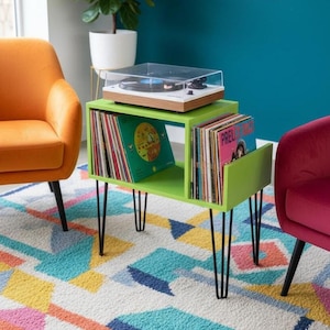 Peut inclure: Un meuble vert clair pour platine vinyle avec des pieds épingles noirs. Le meuble contient des disques vinyles et une platine avec un couvercle transparent. Le meuble est dans une pièce avec des chaises colorées et un tapis à motifs.