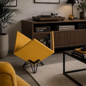 Peut inclure: Un porte-disques vinyle en forme de V jaune moutarde avec un design géométrique et des pieds en épingle en métal noir. Le support est composé de panneaux inclinés créant un espace de rangement central pour plusieurs albums vinyles.