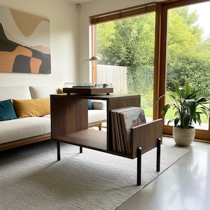Könnte beinhalten: Eine Schallplattenspieler-Konsole aus dunklem Holz mit einem rechteckigen, offenen Staufach für Vinylschallplatten, einer flachen Oberseite für einen Plattenspieler und vier schlanken, schwarzen Metallbeinen in einem schlichten Design.