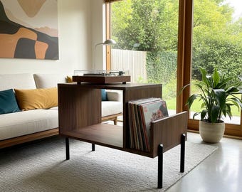 May include: A dark wood record player console featuring a rectangular open shelf design for vinyl storage, supported by four slender black metal legs. A turntable with a clear dust cover sits on the top surface, while a collection of vinyl records is organized vertically in the side compartment.