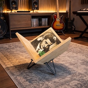 Può includere: Un contenitore in legno chiaro per dischi in vinile dal design a forma di V, sostenuto da quattro sottili gambe a forcina in metallo nero. Il contenitore ospita un singolo disco in vinile con un ritratto in bianco e nero di un uomo.