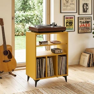 Può includere: Un mobiletto porta giradischi in legno color giallo senape con quattro gambe nere affusolate. L'unità presenta una superficie superiore aperta per il giradischi, due ripiani orizzontali e tre scomparti verticali per i dischi in vinile.