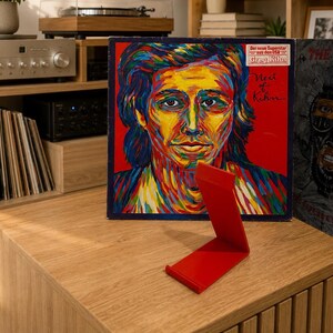Op de afbeelding: Een felrode, L-vormige metalen standaard voor het uitstallen van voorwerpen, geplaatst voor een vinylplatenhoes met de titel Next of Kihn van Greg Kihn. De hoes toont een kleurrijk, impressionistisch portret van een persoon tegen een rode achtergrond.