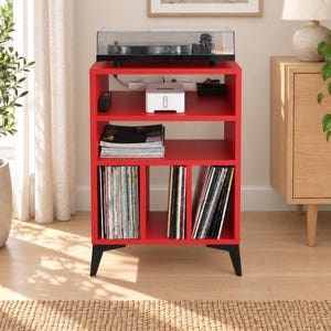 May include: A bright red wooden media console with a black turntable on top. The unit features an open middle shelf holding a white electronic device and a stack of magazines, plus three lower vertical compartments filled with vinyl records.