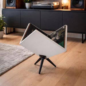 May include: A white vinyl record holder with black legs, displaying two album covers. The holder is angled, showcasing the records. The background includes a black cabinet, speakers, and a turntable, suggesting a music-themed setting.