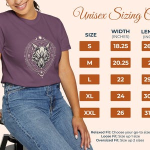 Pu&ograve; includere: Una maglietta viola con un disegno geometrico di una testa di lupo all'interno di un cerchio. L'immagine include una tabella delle taglie con misure in centimetri e il testo "Unisex Sizing Chart". La modella indossa jeans blu.