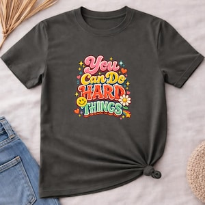May include: A dark gray short-sleeve t-shirt featuring a colorful graphic print on the chest. The text reads You Can Do HARD THINGS in a multi-colored, layered font, surrounded by small decorative elements including stars, hearts, a smiley face, and a flower.
