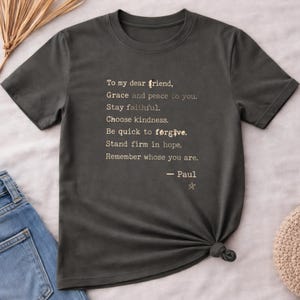 May include: A dark charcoal gray short-sleeved t-shirt featuring white typewriter-style text that reads: To my dear friend, Grace and peace to you. Stay faithful. Choose kindness. Be quick to forgive. Stand firm in hope. Remember whose you are. - Paul.