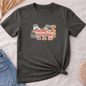 May include: A dark gray short-sleeved t-shirt featuring a graphic with the text "The shirt is decodable" in a retro font. The design includes illustrations of a stack of books, a pencil, and various educational labels with arrows pointing to words.