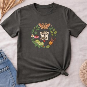 May include: A charcoal gray t-shirt featuring a circular graphic of a moth, a caterpillar, a beetle, and a ladybug surrounded by leaves and flowers. The text "BUGS NEED HUGS" is printed in the center with a small red heart below the words.