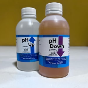 May include: Two plastic bottles with white lids. One is clear, labelled "pH Up" with a blue arrow. The other contains a light brown liquid, labelled "pH Down" with a purple arrow. Both bottles have blue and white labels.