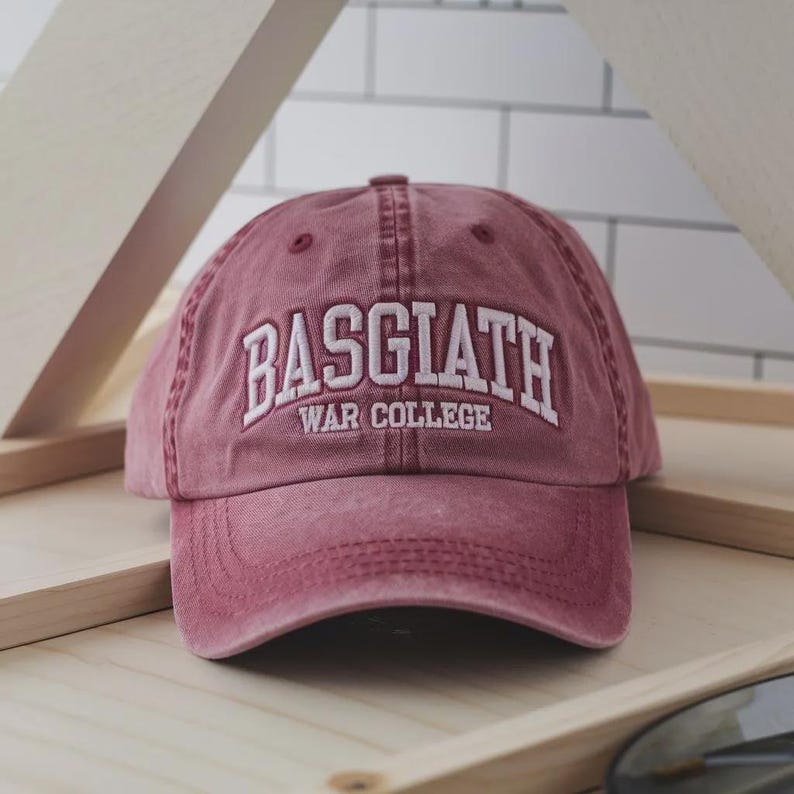 Op de afbeelding: Een bordeauxrode baseballcap met de tekst "BASGIATH WAR COLLEGE" in witte letters geborduurd. De cap heeft een gebogen klep en een verweerde uitstraling. De hoed is gemaakt van een zachte stof.