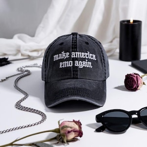 May include: A black, distressed baseball cap with the embroidered phrase "make america emo again" in white. The cap is the focal point, with a silver chain, sunglasses, and a candle in the background. A dark rose is also visible.