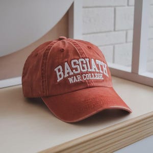 Op de afbeelding: Een roestkleurige baseballcap met de witte tekst "BASGIATH WAR COLLEGE". De pet, gemaakt van een zacht, versleten materiaal, ligt op een houten oppervlak. De hoed is een klassiek model met een gebogen klep.