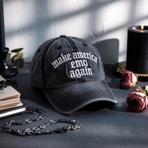 May include: A dark gray baseball cap with the phrase "make america emo again" embroidered in white. The cap is made of a worn fabric and has a curved brim. The cap is on a dark surface.