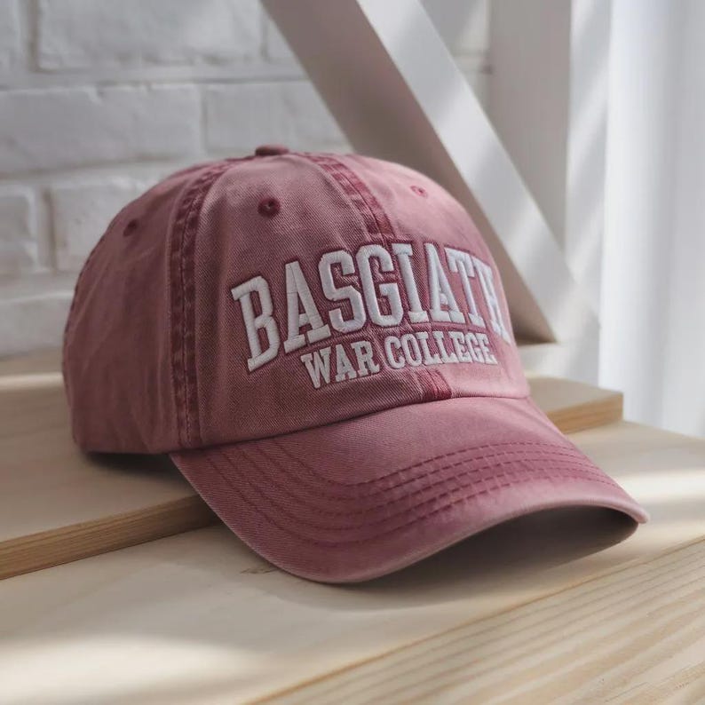 Op de afbeelding: Stofroze baseballpet met de tekst "BASGIAT WAR COLLEGE" in het wit geborduurd. De pet is gemaakt van een zacht, versleten materiaal en ligt op een houten oppervlak. De hoed heeft een gebogen klep en een klassiek zes-panelen ontwerp.