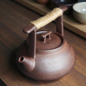 Puede incluir: Una tetera de cerámica marrón con cuerpo redondeado y boquilla. El asa es de cerámica marrón envuelta con cuerda de color natural. La tapa tiene un asa pequeña. La tetera está diseñada para preparar té.