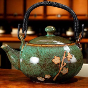 May include: A ceramic teapot with a green and brown glaze, featuring a black woven handle and floral accents. The teapot has a rounded shape with a spout and a lid with a small knob. The floral design includes light brown flowers and stems.