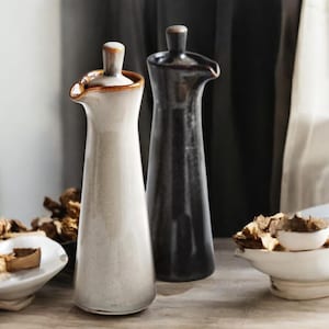 May include: Two ceramic oil and vinegar bottles. One is off-white with a brown rim and a wooden stopper. The other is black. Both have a slender, tapered shape and a spout for pouring. They are on a wooden surface.