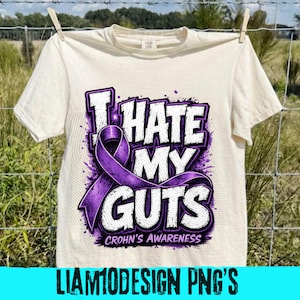 Op de afbeelding: Crèmekleurig T-shirt met de tekst "I HATE MY GUTS" in wit en paars, met een paars lint en de woorden "CROHN'S AWARENESS". Het shirt hangt aan een waslijn. De onderkant van het shirt heeft de tekst "LIAM10DESIGN PNG'S" in een blauwe rechthoek.