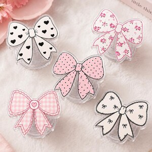 May include: Five decorative bow-shaped items featuring various patterns: one white with black hearts, one white with pink floral prints, one pink with black polka dots, one pink gingham check with a central heart, and one white with black bow outlines.