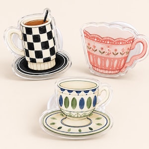 May include: Three acrylic decorative pieces shaped like teacups and saucers. One features a black and white checkered mug with a black saucer. Another is a pink teacup with floral details. The third is a cream teacup with blue and green oval patterns.