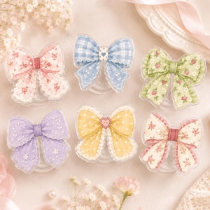 May include: Six decorative bow-shaped phone grips featuring various patterns including pink floral, blue gingham, green strawberry, purple floral, yellow polka dot with a heart button, and cream floral with red accents, all with scalloped edges.